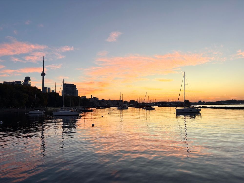 Sunrise over a marina on the Toronto waterfront. Pink, yellow, and orange light is reflected in the calm water where a few boats are moored. 