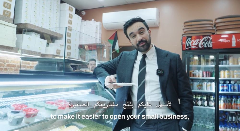 Still from a video featuring Zohran Mamdani standing against a counter in a take away shop, holding a paper plate with knafeh. Text in arabic and english reads “to make it easier to open your small business”. 