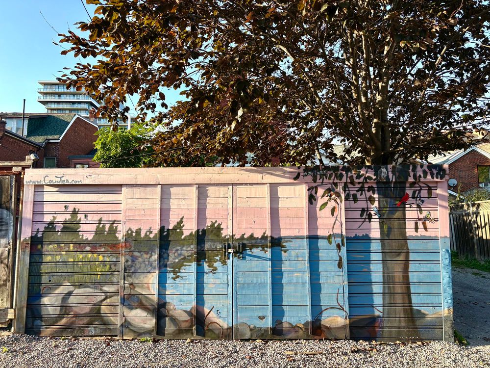 Mural on a garage door in a laneway depicting a bay of water on a lake with a tree-lined, rocky shoreline and pink sky overhead. A large tree painted in the foreground aligns with the top of a real tree growing behind the garage. Art by Colin Tea.