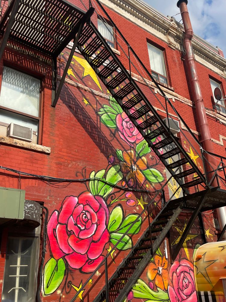 Large mural of red flowers and stars on the side of a red brick building, tucked behind a metal fire escape. 