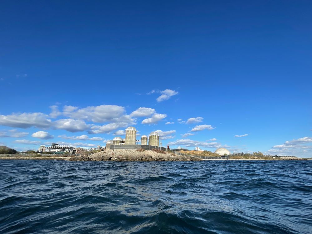 The West Island at Ontario place viewed from the water. An empty space once filled with trees on a rocky shoreline on Toronto’s waterfront. Black fencing surrounds the perimeter.