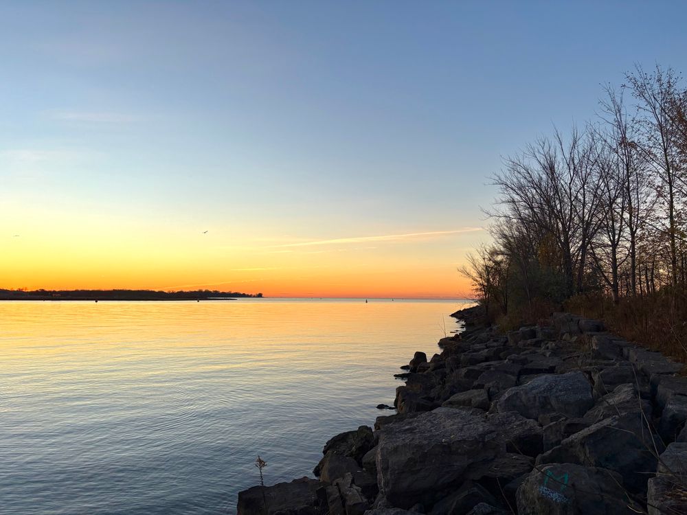 Sunrise over Lake Ontario in a Toronto waterfront park. Rocky tree-lined shoreline and the Toronto islands visible in the distance. Clear blue sky overhead.