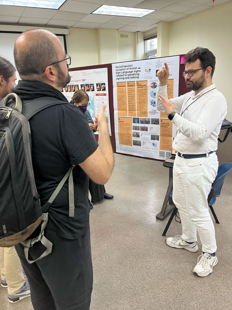 Me, a man dressed in white shirt and trousers standing next to my poster “Sociolinguistic variation of Polish Sign Language signs related to "speaking", "signing"”. I’m explaining the contents of the poster in International Sign to a couple of people standing in front of me.