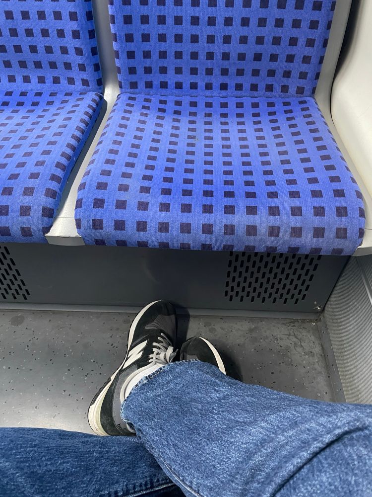 Bild überkreuzter Beine mit Blick Richtung Boden auf Turnschuhe in einem sbahn-Abteil. Die Person trägt eine blaue Jeans, weise Socken und grau-schwarze, ausgewaschene Turnschuhe der Marke new balance. Die beiden gegenüberliegenden Sitze sind leer. 