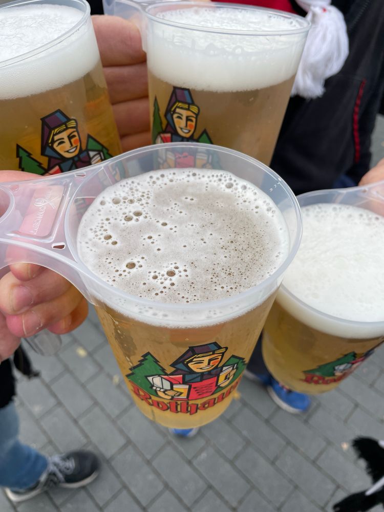 Vier volle Becher mit Rothauspils vor dem Station vom SC Freiburg beim Spiel gegen Eintracht Frankfurt. Die Personen halten die Becher im Kreis für eine Detailaufnahme. Man sieht keine Personen, lediglich den grau gepflasterten Boden und Schuhe leicht unscharf.