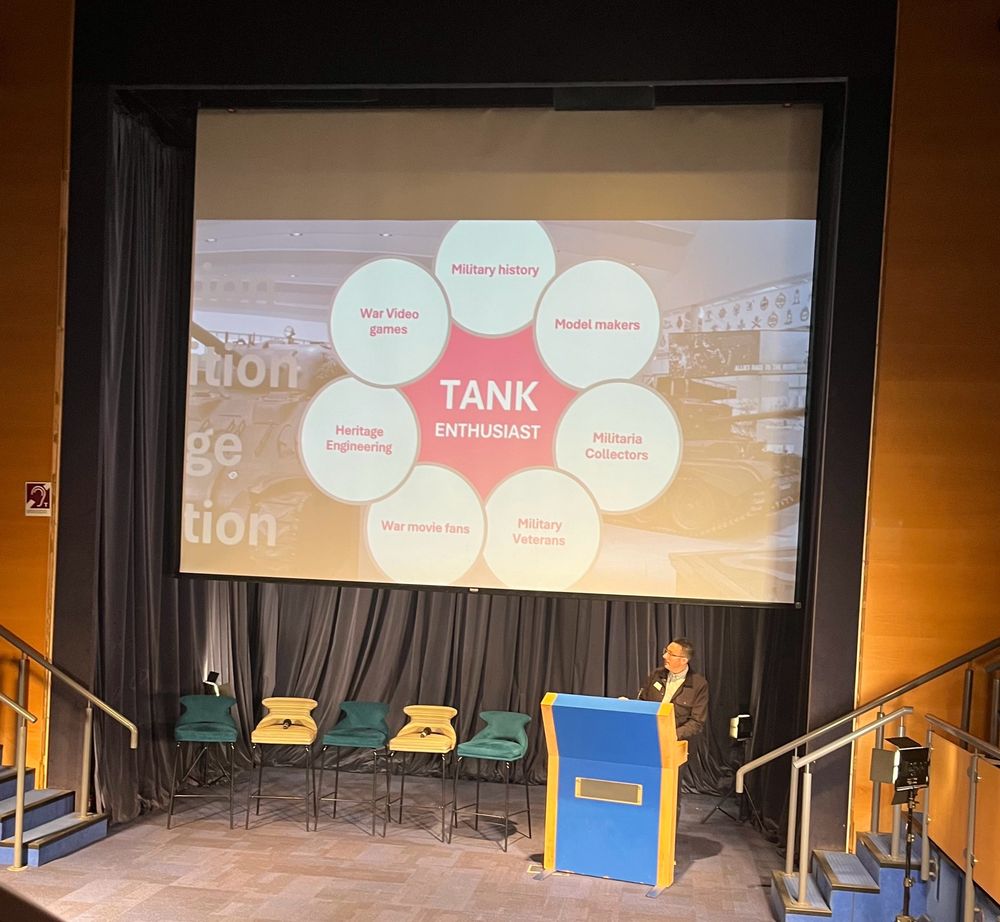 Photo of a presenter standing below their presentation showing a circular diagram of many tank museum audience types including: war video games, military history, heritage engineering, war movie fans, military veterans, model makers and militaria collectors