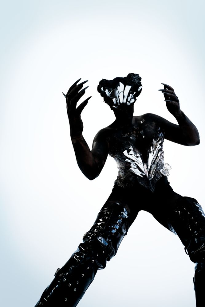 Photograph of a model dressed in a costume resembljng a shadowlike creature. The face and body seem to consist of shattered mirrors, and the model is wearing long leather overknee boots, long nails and black special effects makeup and bodypaint. The photo of the black silhouette ist taken on a white, contrasting background. 