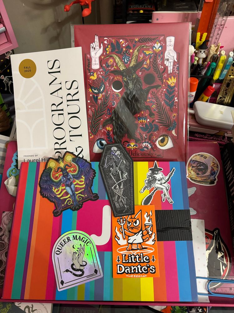 A colorful desk full of fun - program for a cemetery, print of black Phillip beautiful decorated in a folk style, a Baphomet sticker, two skeletons sticker, Queer Magic Sticker, a Witch in a broom and a Little Dante Sticker
