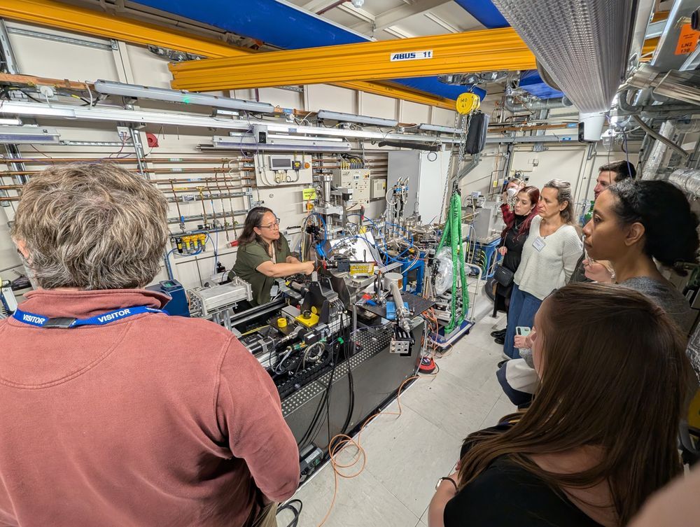 Visitors of the Harwell Heritage Network meeting observes Dr. Susan Nehzati explaining a data collection setup inside a beamline laboratory. The room is filled with scientific equipment, metal piping, cables, and control systems.