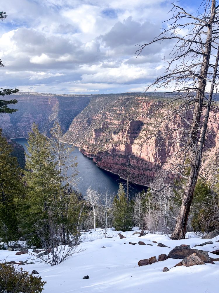 photo of Flaming Gorge in the late afternoon, snow and trees in the foreground and a river flowing through the gorge behind it, the sky is blue with fluffy white clouds