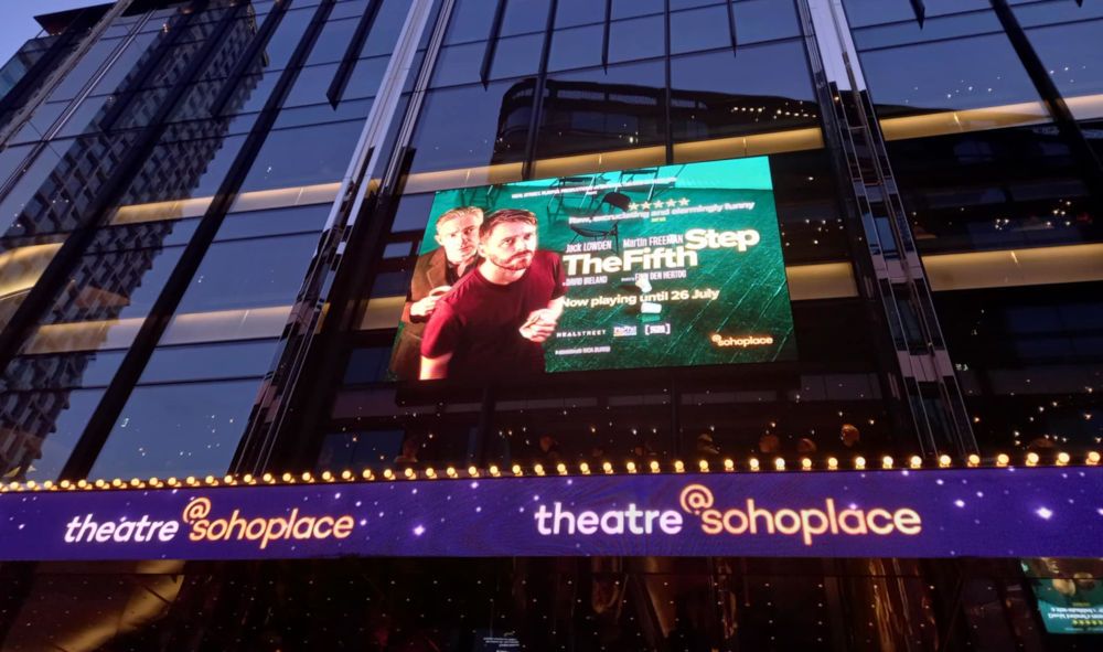 Illuminated screen showing an advert for The Fifth Step, with Martin Freeman and Jack Lowden looking upwards. It sits on a glass and steel building above a digital, blue hoarding for theatre @ sohoplace. Evening.