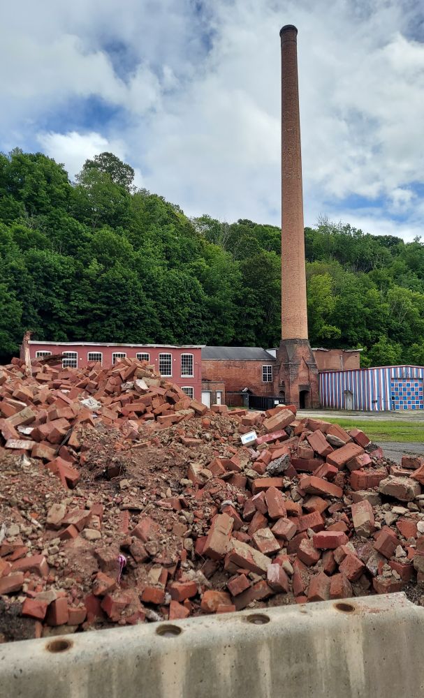 In the foreground is a large pile of broken bricks and a cement barricade. In the background is a brick building and garage with red white and blue stripes. In between those is an incredibly tall smoke chimney that looks like it's seen better days. 