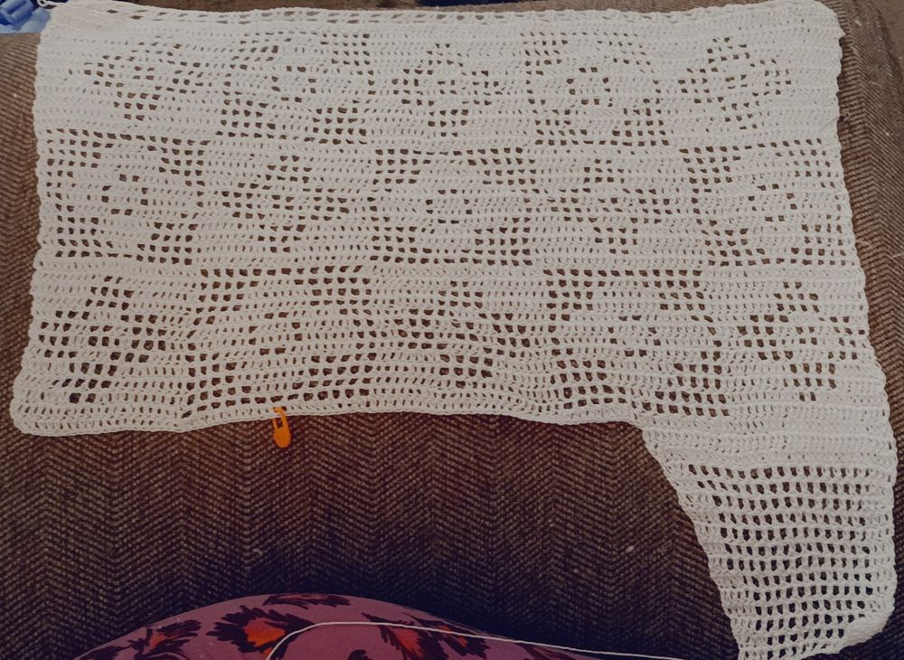 A filet crochet piece in progress; mostly showcasing a retro flower grid design and one side has the beginning of a mesh tie extending forward. 