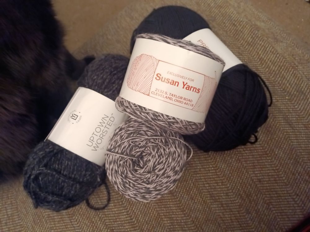 A pile of yarns; two small cakes of marled pink and gray sock yarn, one heather charcoal worsted weight skein, and some solid black yarn. 