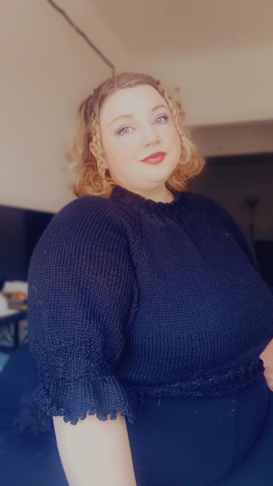 Morgan, a blonde plus size woman, sits posed to show a black hand- knit raglan blouse. It has twisted cable details at the collar and hem, and puffed poet sleeves with a ruffle and picot edge. Her hair is braided at the front and partially pulled back, and she has a serene, pleasant expression on her face. 