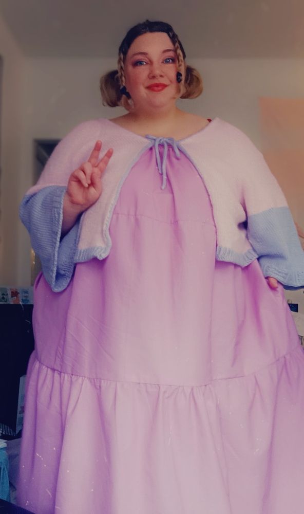Morgan stands, holding up a peace sign. She is wearing a long, 3-tier maxi dress she made, and a hand knit,  pink and purple color block cardigan on top. The dress is a light orchid color, and looks flowy and comfortable. Morgan is smiling at the camera