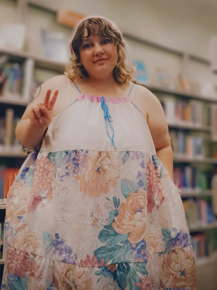 Morgan stands holding a peace sign in front of a blurry bookshelf backdrop. She wears the color blocked summer dress she made- the top is cream, and the skirt is a muted floral print. The straps and ties in the front are bright blue
