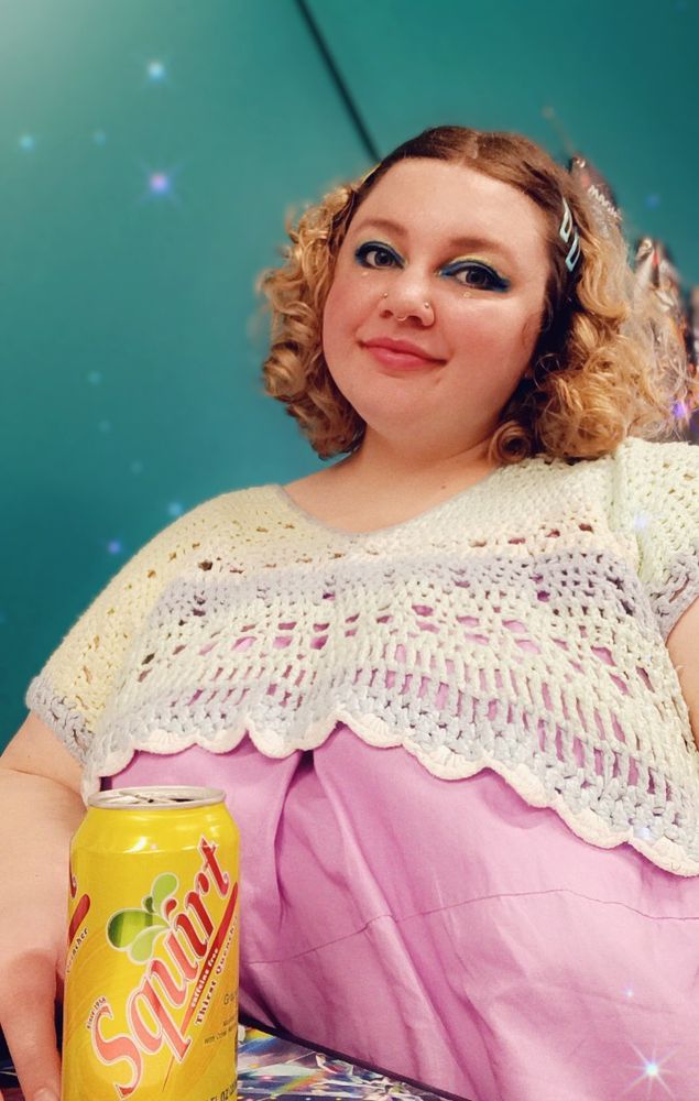 Morgan sits in front of a teal backdrop. Her hair is curly and clipped back, and she wears colorful makeup and a colorful crochet top layered over a pink dress. In the foreground, she holds a yellow-green can of lemon lime soda emblazoned with the word "Squirt" in red. 