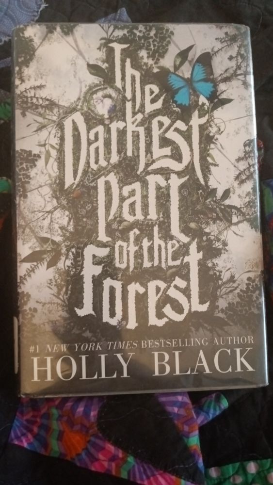 A photo of a hardcover library edition of The Darkest Part of the Forest by Holly Black. The cover features a stark, graphic image of branches and evergreen, with a bright blue butterfly by the text for visual contrast. The book is laid atop a dark and colorful homemade quilt