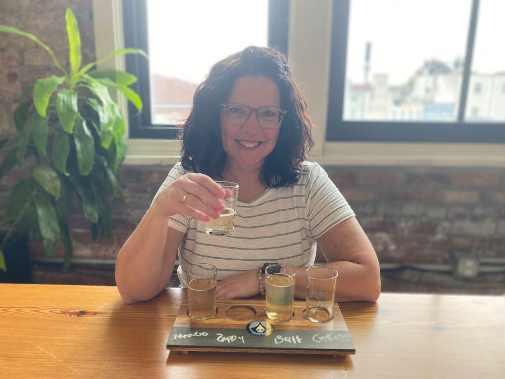 A retired woman in a striped shirt with brown curly hair and glasses sits behind a flight of 4 beers at Rhinegeist Brewery in Cincinnati. 