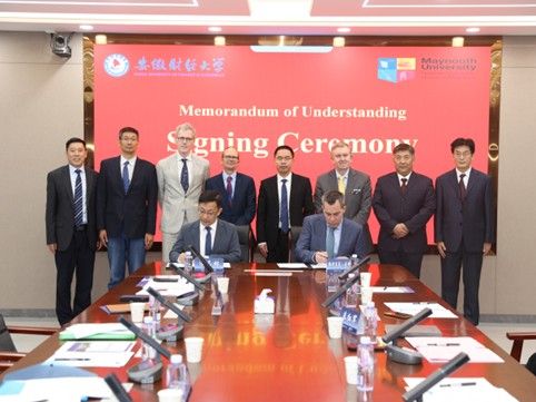A group of men in formal attire are posing in front of a large red banner reading "Memorandum of Understanding Signing Ceremony." Two men are seated at a table in the foreground, with one signing a document. The table is covered with microphones, documents, and water bottles.