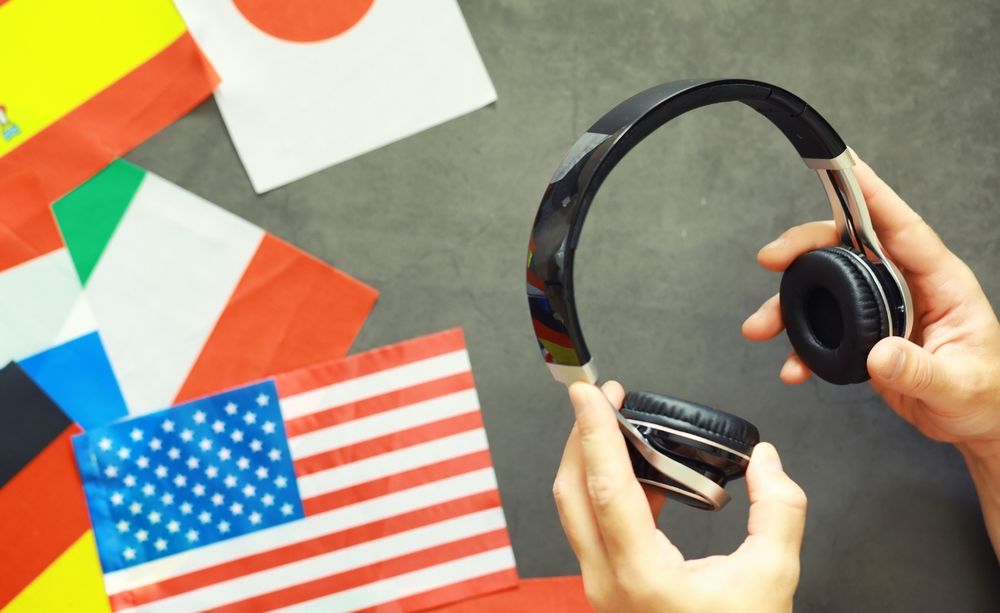 Hands holding black headphones over a table with flags from various countries, including the United States, Japan, and Italy, spread out in the background.