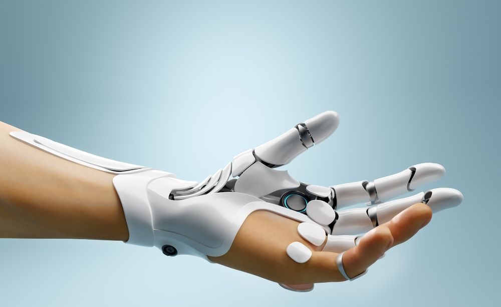 A white robotic hand with sleek, futuristic design extends outward, palm facing up against a soft blue background. The mechanical joints and fingers are detailed, combining metallic and smooth surfaces. The wrist is seamlessly connected to a person's forearm, blending technology and human form.