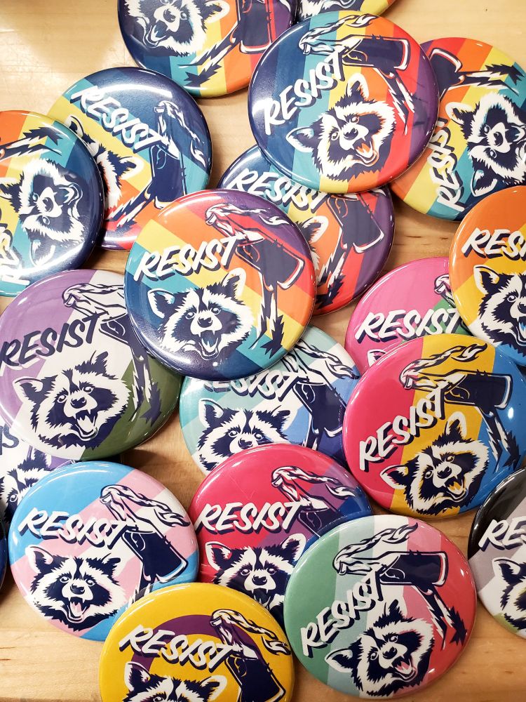 A lot of rainbow and pride flag 2.25" pinback buttons featuring a raccoon throwing a Molotov and the word RESIST