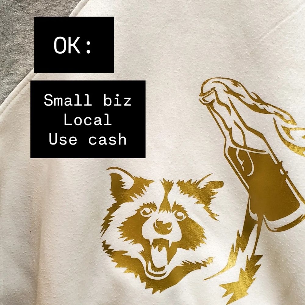 A sweatshirt printed with a raccoon throwing a Molotov design. Text "OK: Small biz, Local, Use Cash"