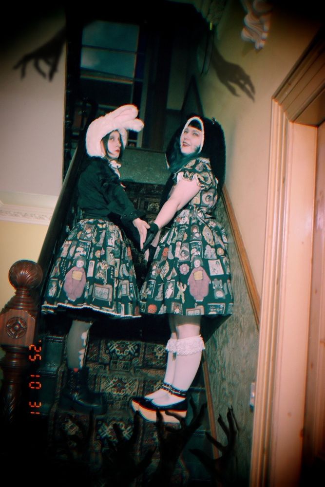 Two gothic lolitas, dressed in Violet Fane and rabbit ear hoods stand in a dark stairwell. Dark shadowy hands reach for them.