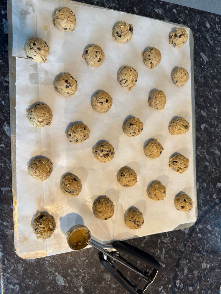A baking tray with perfectly even cookie dough balls ready for baking. Chocolate chip cookies. A cookie dough/ice cream scoop that was used to make the perfectly even cookies is resting on the tray. 