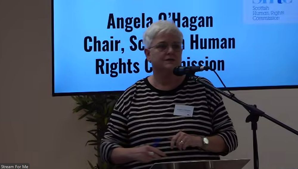 Angela O'Hagan, chair of the Scottish Human Rights Commission, speaks in front of a microphone at the inaugural event of Tax Justice Scotland (8 October 2025), with her name and role projected against a blue background behind her