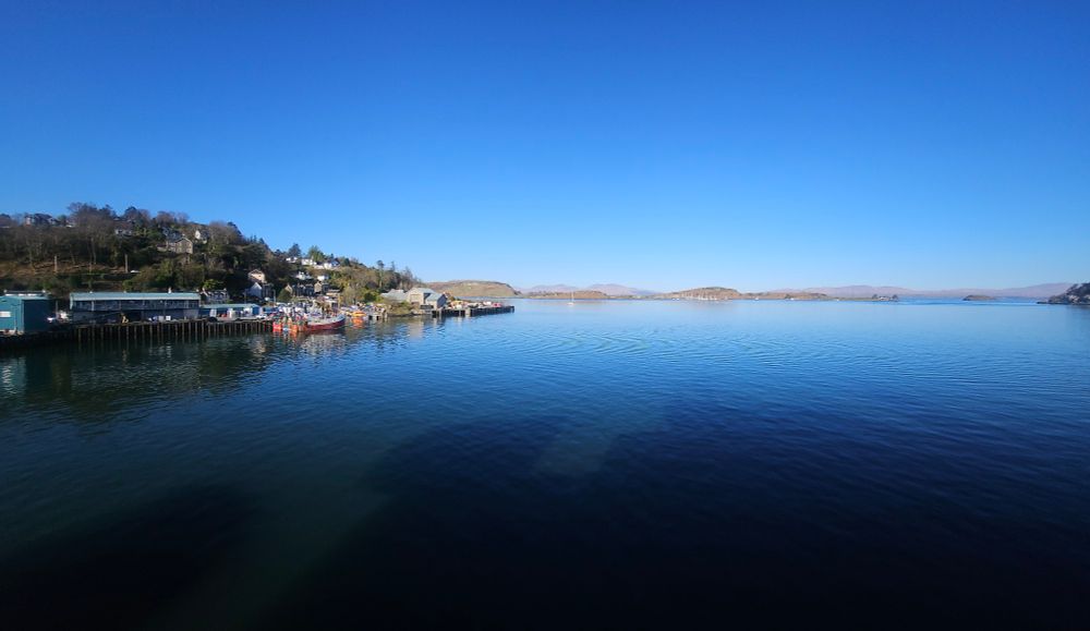 A picture of the harbour in Oban looking out to sea. The headland is on the left and there are islands in the distance. Clear blue sky above