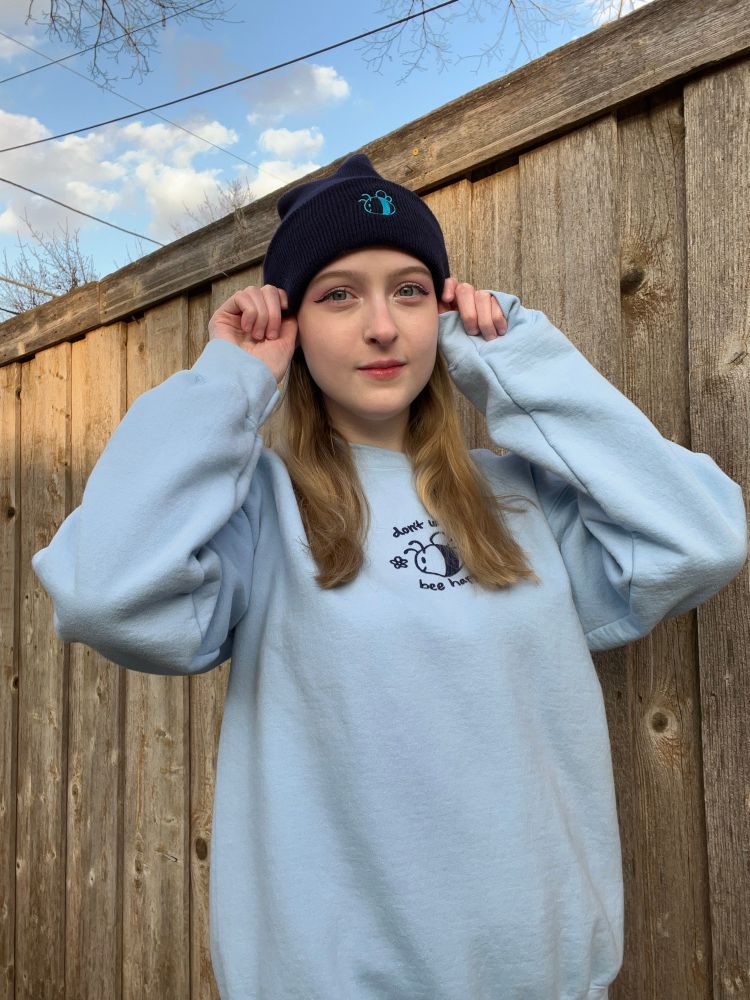 Abbey is wearing a don’t worry bee happy embroidered blue sweatshirt while holding down a dark blue beanie on her head that has a blue embroidered bee