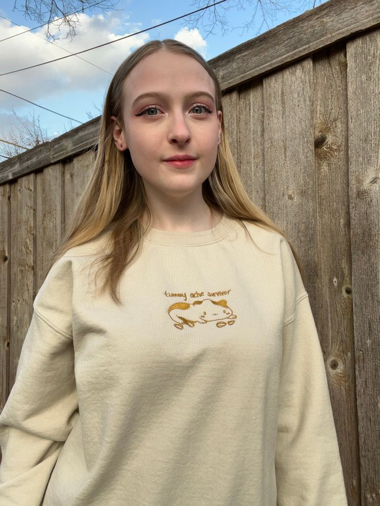 Abbey is wearing a pale yellow sweatshirt that is embroidered with a hamster and it says tummy ache survivor