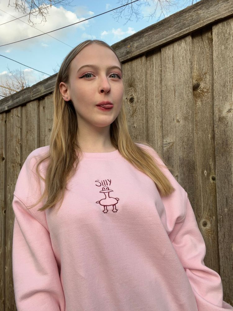 Abbey is wearing a pale pink sweatshirt embroidered with the words “silly” and a goose
