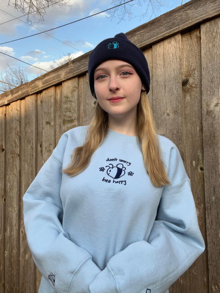 Abbey is wearing a blue sweatshirt that is embroidered with a bee and says don’t worry bee happy