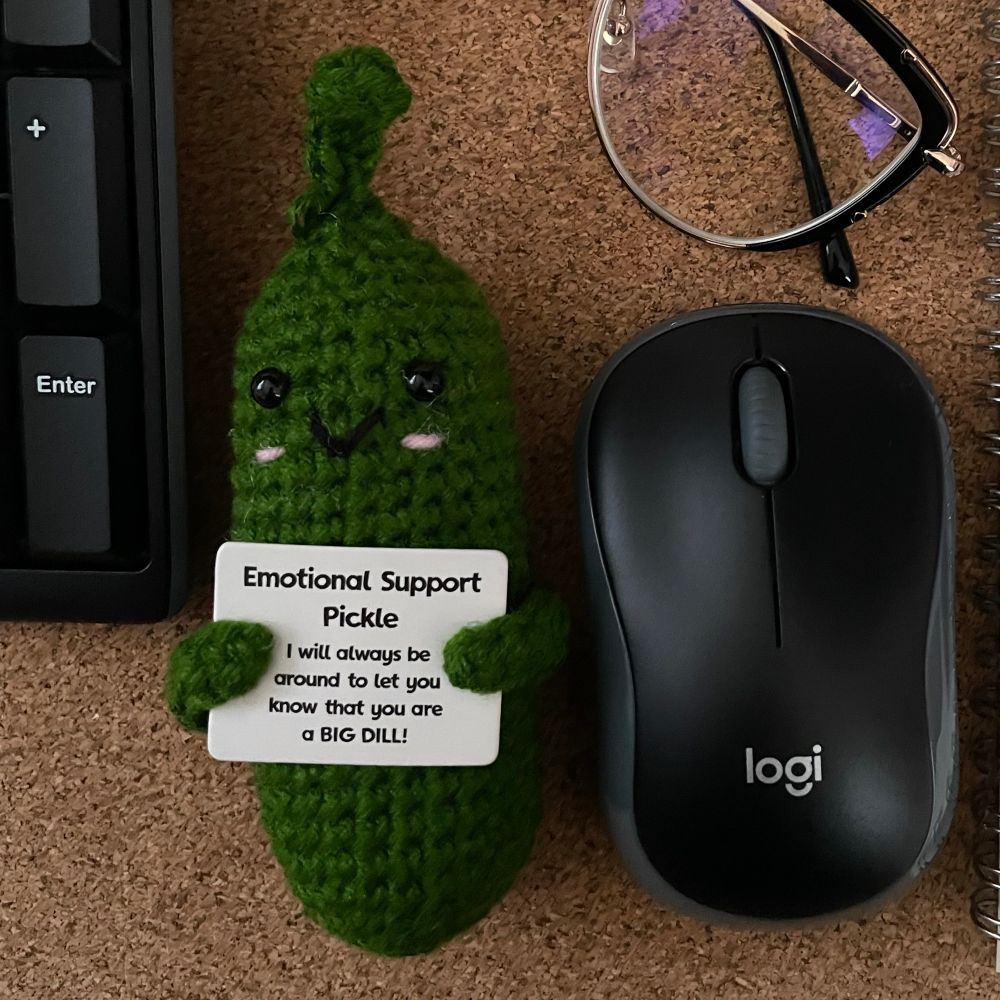 Photo of a crocheted green pickle with cute facial features and little arms holding a sign that says “Emotional Support Pickle - I will always be around to let you know that you are a BIG DILL!” on a cork surface with part of a pair of black and gold rimmed eyeglasses, the edge of a keyboard and a wireless mouse for scale comparison. 