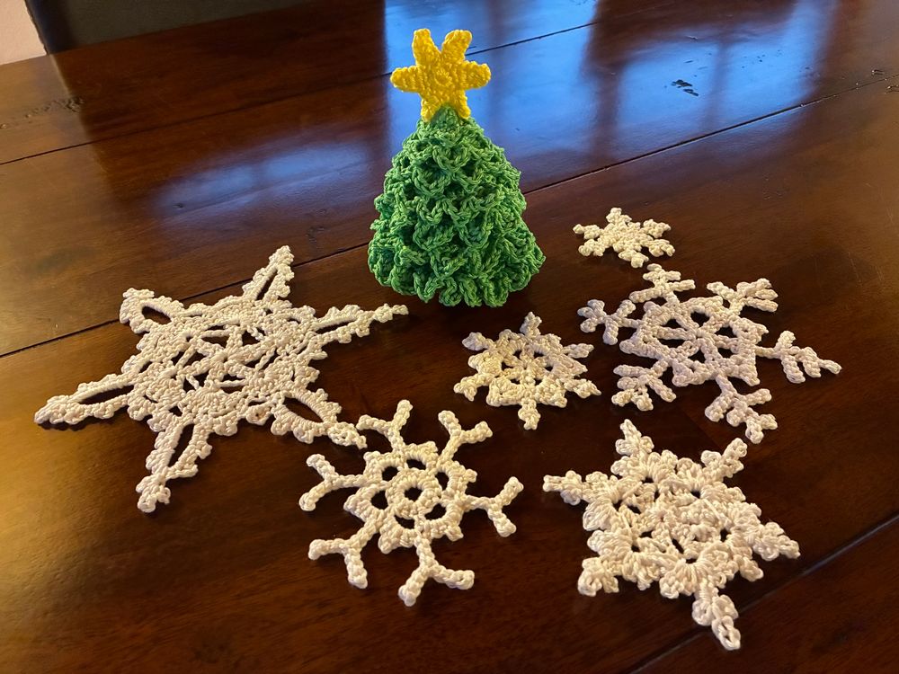 Crocheted twodimensional snowflakes in different sizes around a crocheted threedimensional Christmas tree which has a yellow star on top. Everything spread out on a dark brown table.