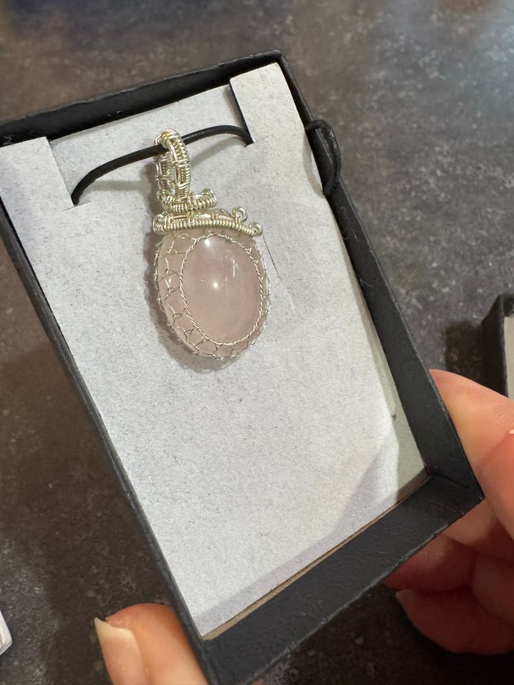 A white person’s fingertips are holding a jewelry box with a beautiful, pale pink quartz pendant. The opaque stone is polished to a high shine and is a very light color. The silver wire wrapping around the stone forms a very fine mesh around the edges and a sturdy loop for the necklace to pass through. 