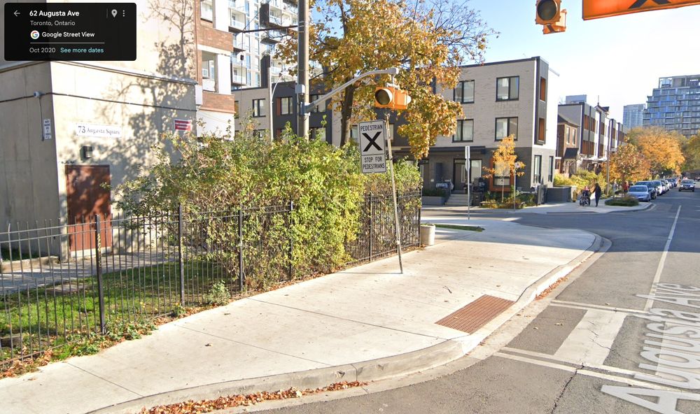 A screenshot of a Google Street View, at August Ave. and Paul Ln Gardens in Toronto, which shows a pedestrian call button at knee height for adults (in the very middle of the screenshot). There is a wide sidewalk with a crosswalk that takes up most of the screenshot.