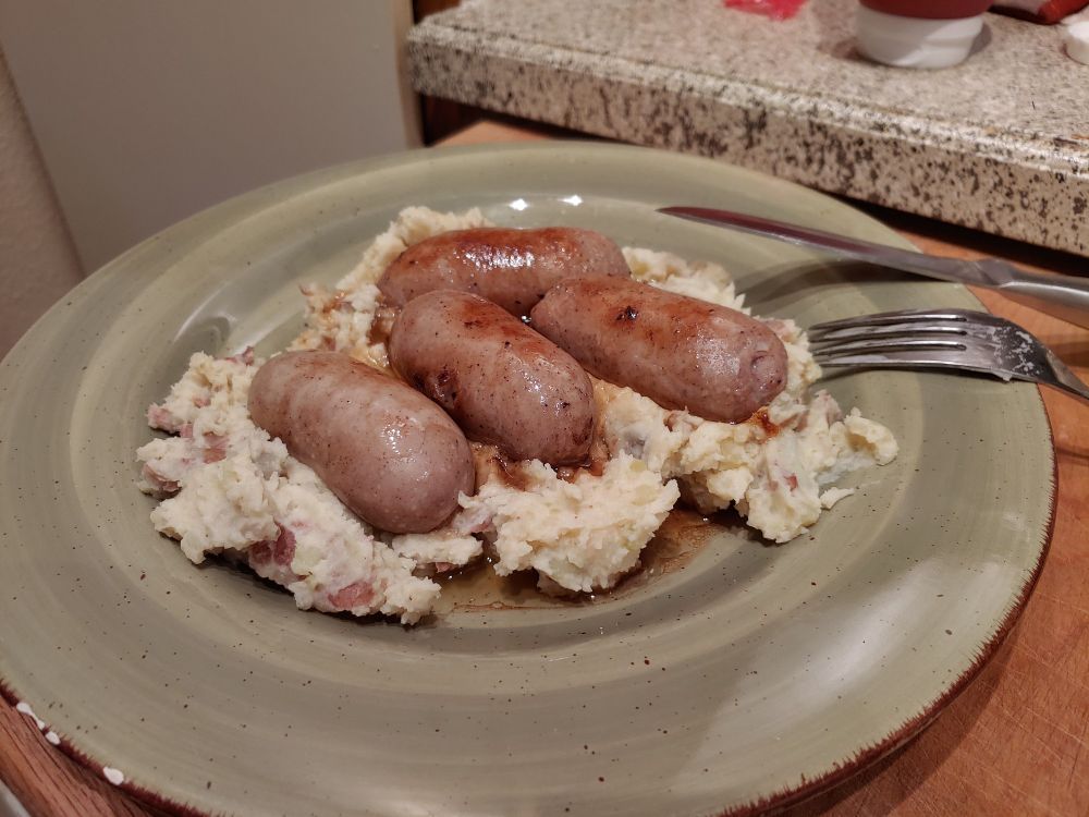 four small banger sausages and greasy gravy on a bed of skins-in mashed potatoes, on a sage green ceramic plate