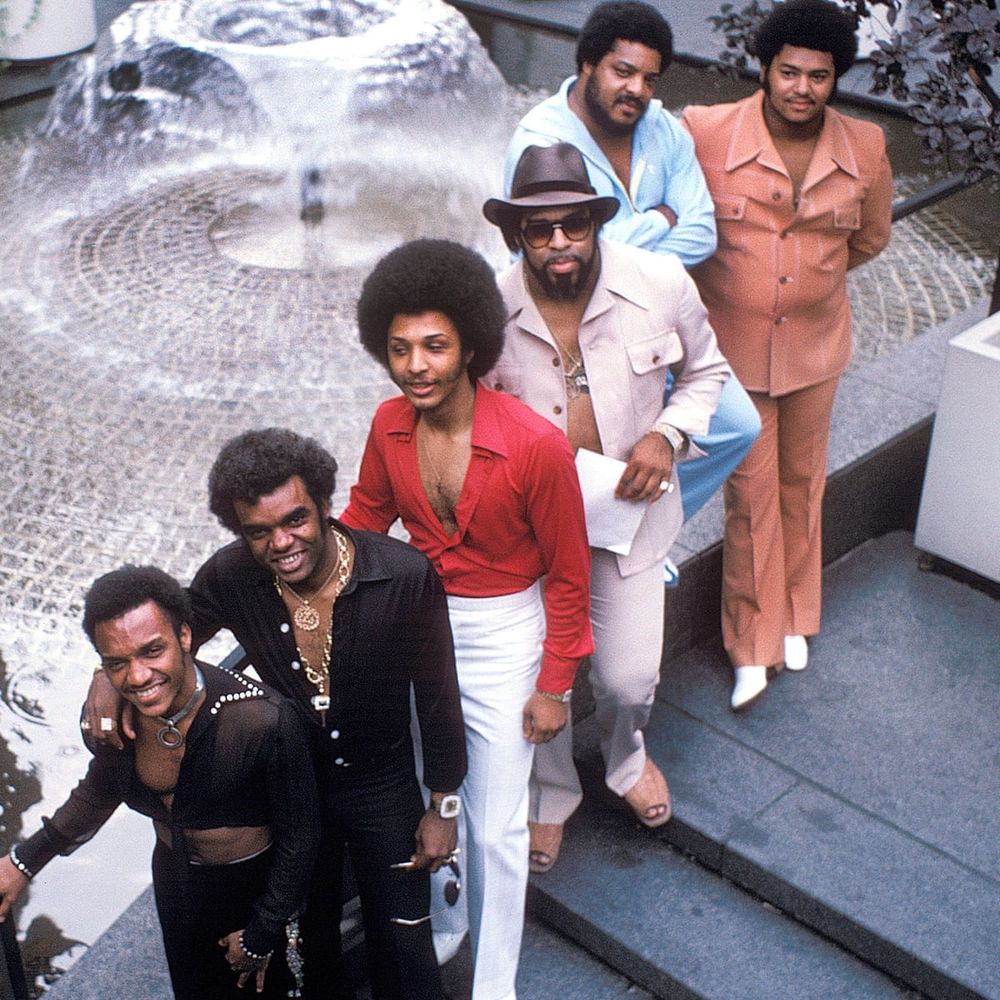 Photo of the 6 members of the Isley Brothers group from above. They are standing outside next to a fountain, smiling at the camera 