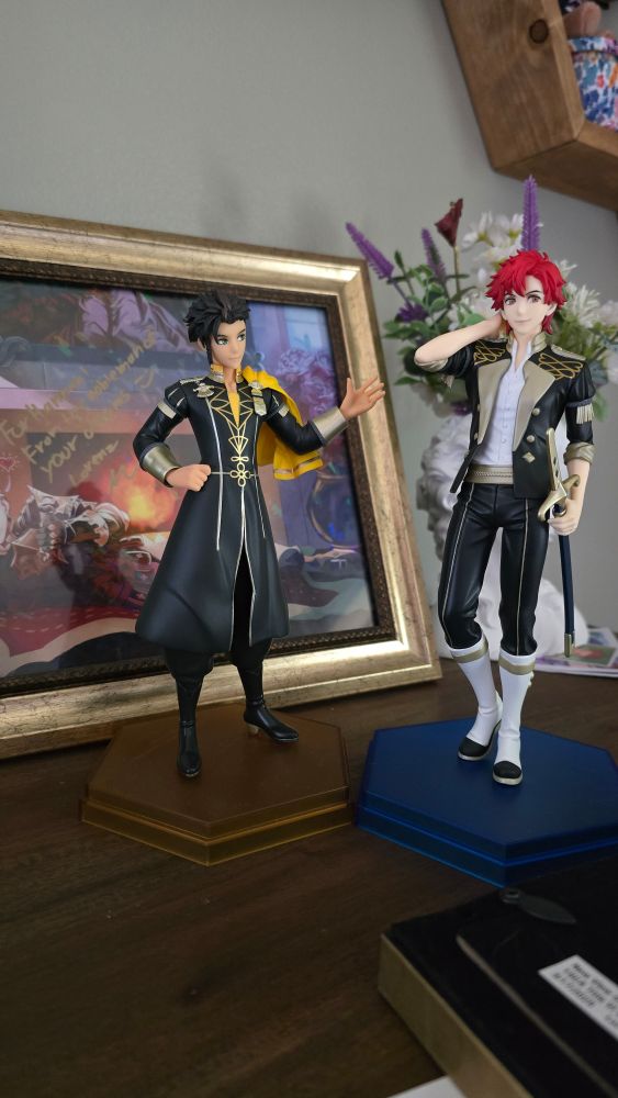 The Claude and Sylvain figures from the game Fire Emblem Three Houses, glancing askance at each other. Claude is short and powerful while Sylvain is lackadaisical snd tall. A photo of Lorenz in the background peeks coyly at them.