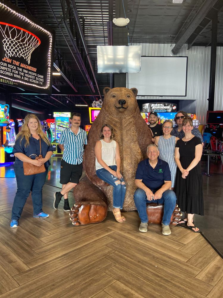 Group photo of Franco Institute staff. Eight people smile and pose around a large bear statue in an arcade setting.
