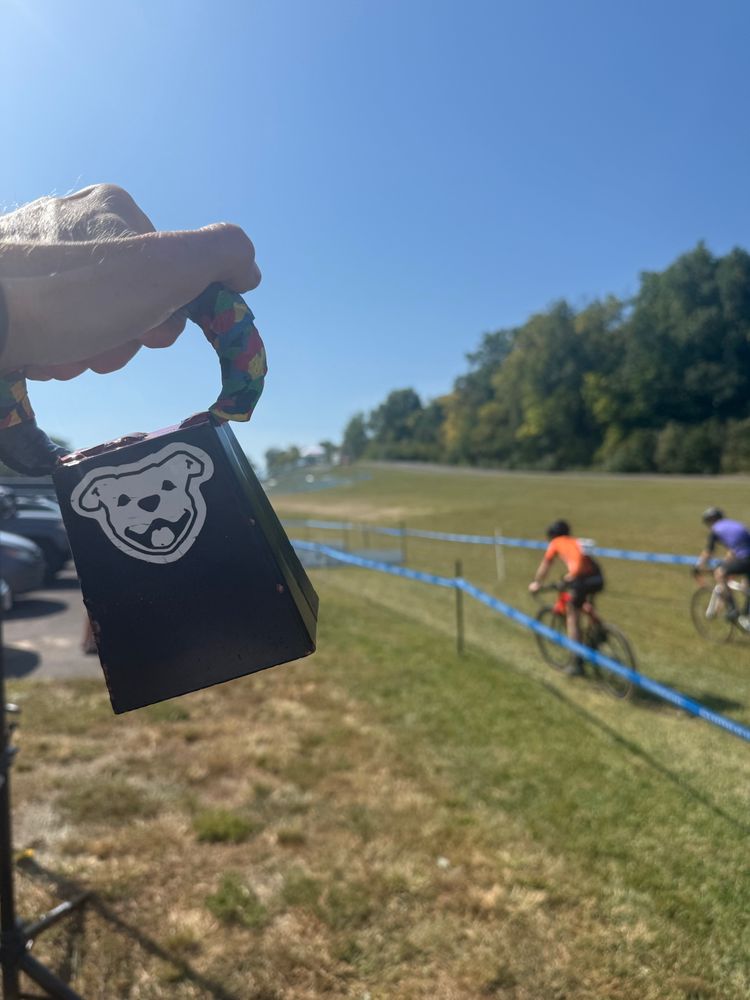 À cowbell in foreground and two cyclocross racer behind course tape in the background 