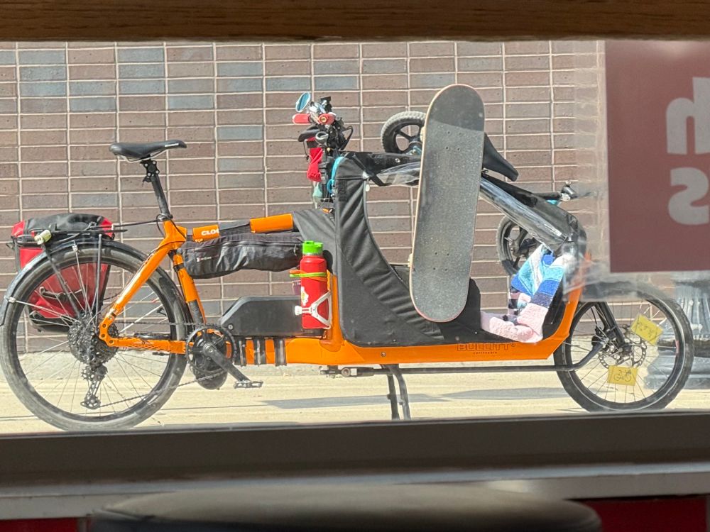 A cargo bike is parked outside a donut shop and the passenger compartment is empty. A skateboard and a balance bike are secured to the sides of the passenger compartment. 
