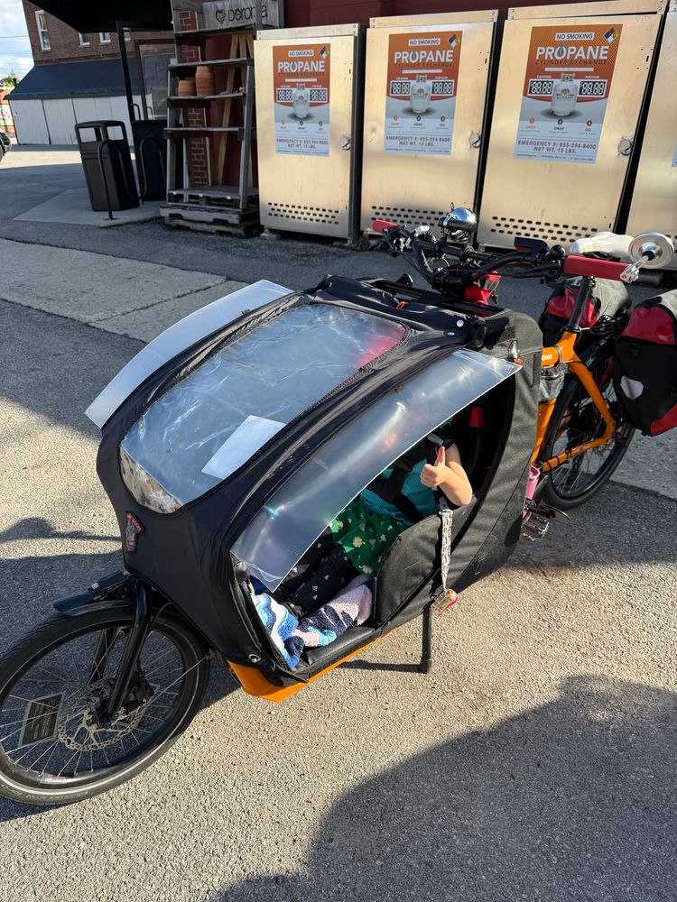 A front loader cargo bike has a window sized piece of plexiglass strapped under the fabric windshield of the passenger capsule, the passengers's hand makes a thumbs up gesture out the side of the capsule 