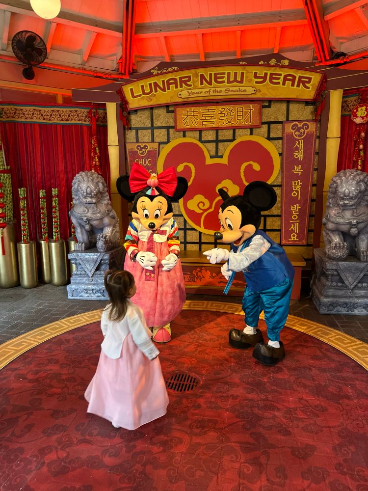 Little girl wearing a light pink hanbok meeting with Minnie Mouse and a Mickey Mouse who are also wearing hanboks. A sign in the back in gold and red says Lunar New Year Year of the Snake.