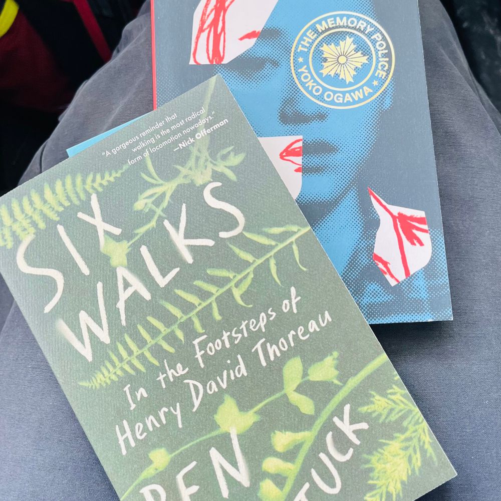 Two books sit on someone’s lap. On top is a green book with images of leaves and  the title Six Walks: In the Footsteps of Henry David Thoreau. The author is Ben Ahattuck but his name is partly cut off in the photo. Underneath is a blue book with a photo of a woman on the cover. Parts of her face are obscured with oddly shaped blobs of pink and red. The book is The Memory Police by Yoko Ogawa. 